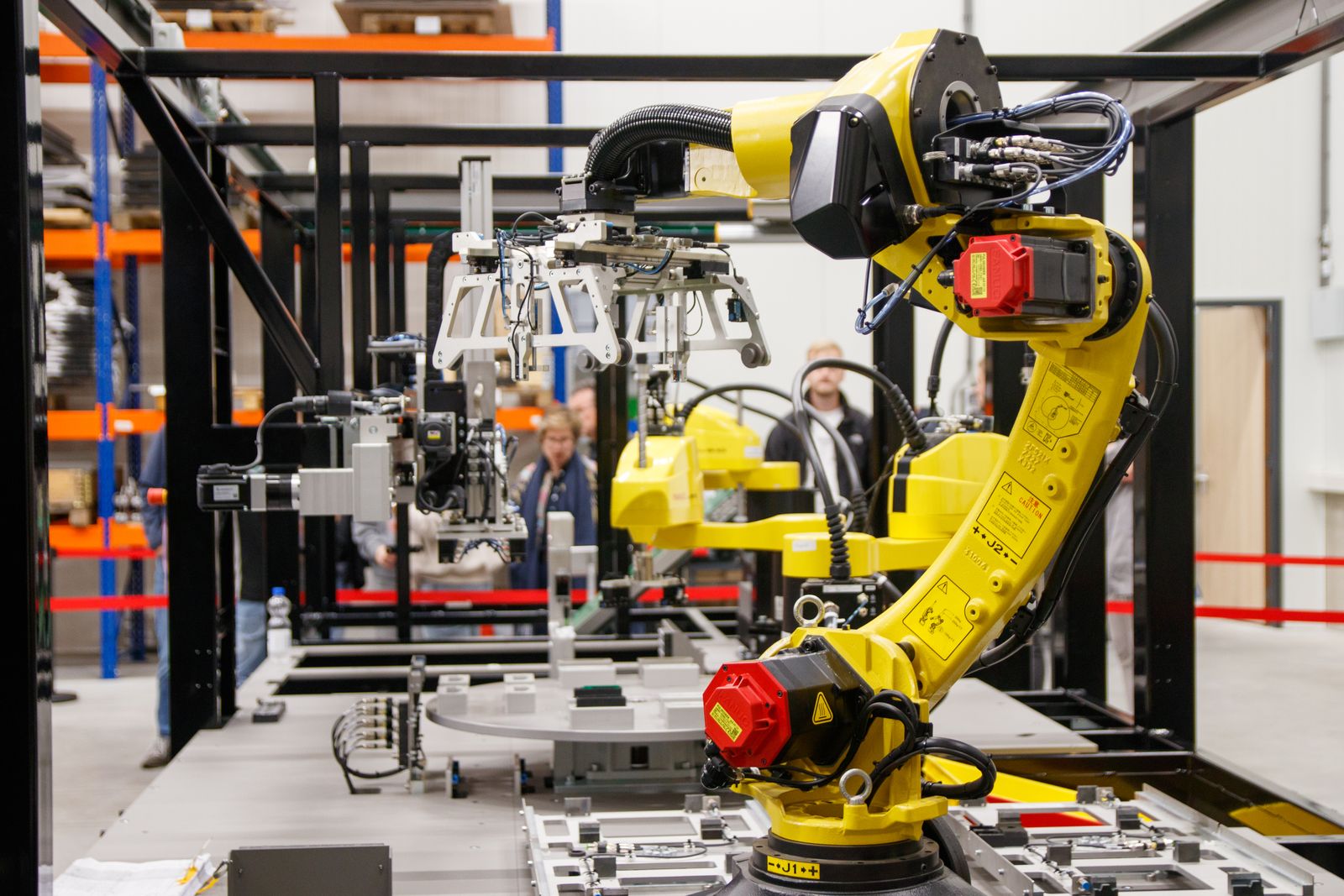 A yellow industrial robot arm on a factory line, its jointed segments and red emergency stop button in focus, two workers blurred in the background behind the safety frame.