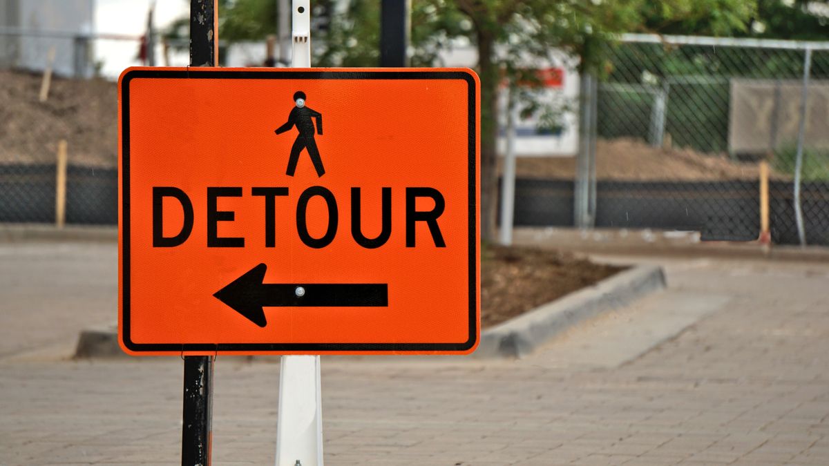 Una señal de tráfico rectangular naranja con la palabra DETOUR, un pictograma negro de peatón encima y una flecha negra que apunta a la izquierda, sujeta a un poste negro delante de una valla metálica desenfocada. Un override hace lo mismo con una dependencia: mismo destino, otra ruta.