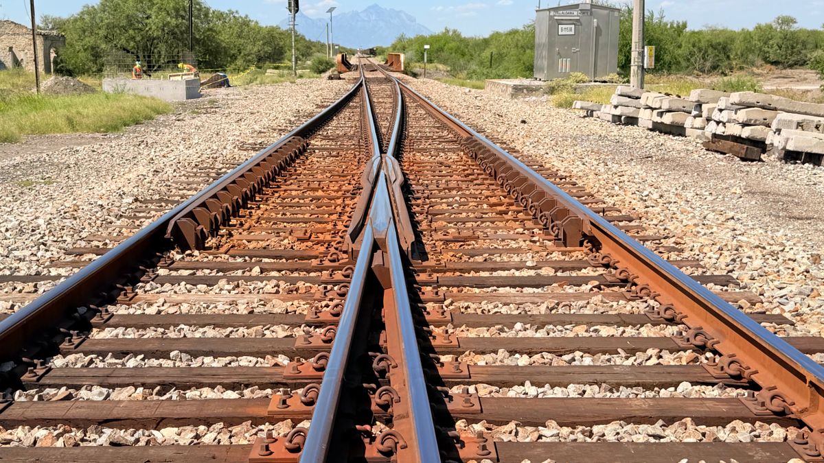 Eine Eisenbahnweiche auf Schienenhöhe fotografiert: zwei rostfarbene Gleise auf Betonschwellen laufen auf hellem Schotter zu einem zusammen, dahinter ein grauer Schaltschrank, gestapelte Betonschwellen und ein Berg am Horizont