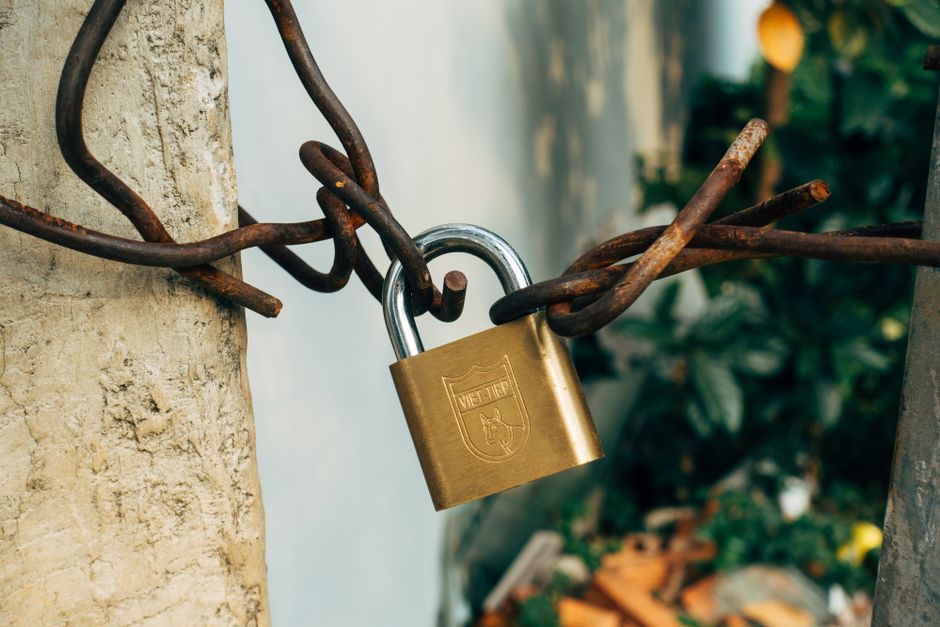 Un cadenas en laiton fermé sur du fil de fer rouillé torsadé, tendu entre deux poteaux en béton, verdure floue à l'arrière-plan. Le manifeste est le dessin ; le lockfile est le moment où quelque chose a été figé.