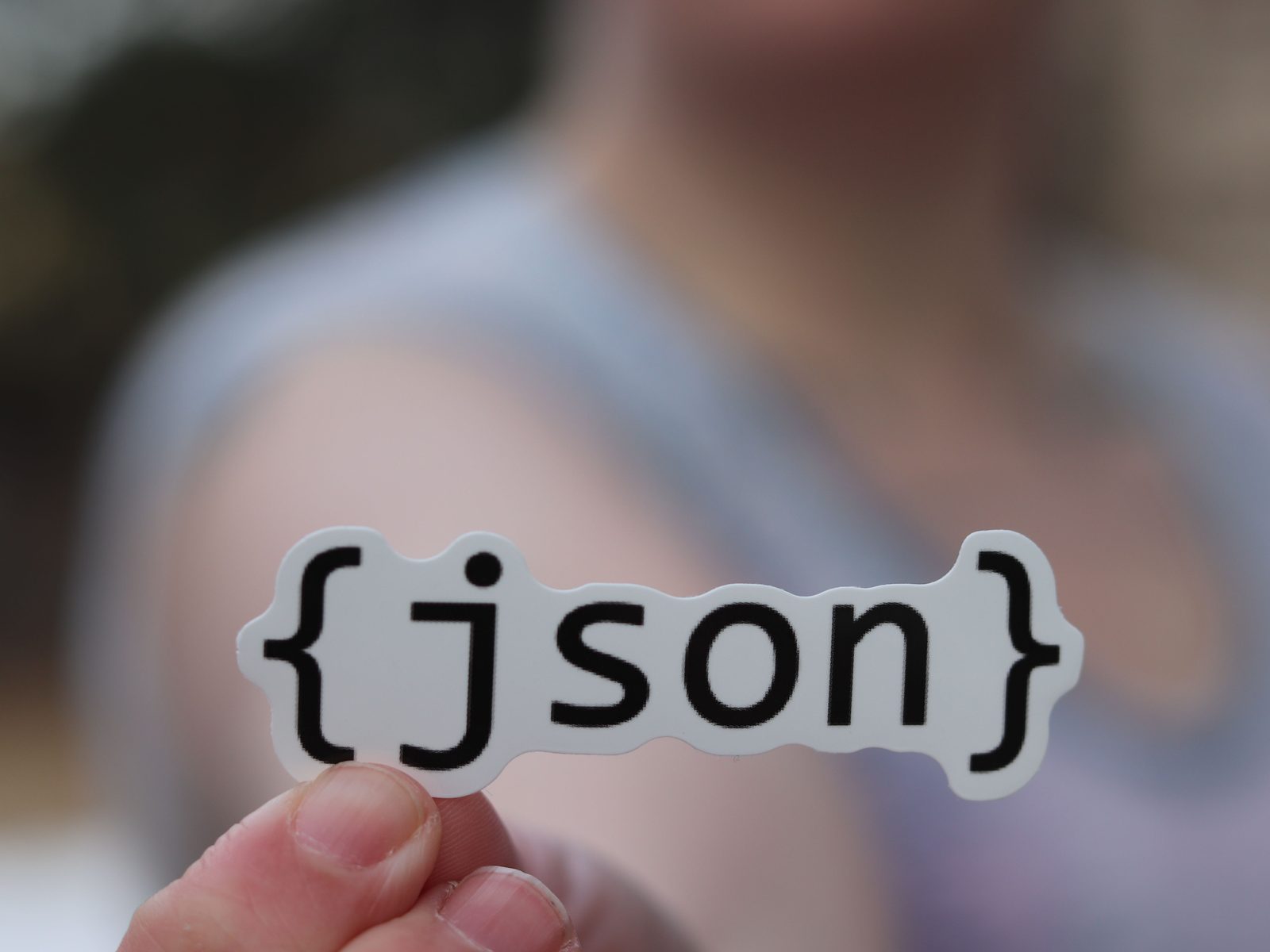 Una mano sosteniendo una pegatina blanca con la palabra json, con una figura desenfocada al fondo