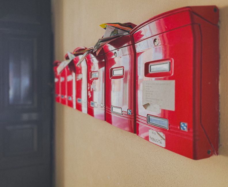 A row of glossy red apartment letterboxes on a beige wall, several stuffed so full that magazines stick out of the top
