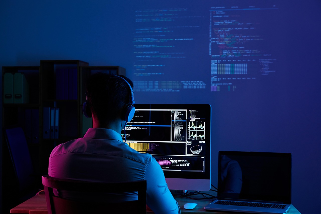 A developer seen from behind, wearing headphones, working at a monitor showing code in a dark blue-lit room