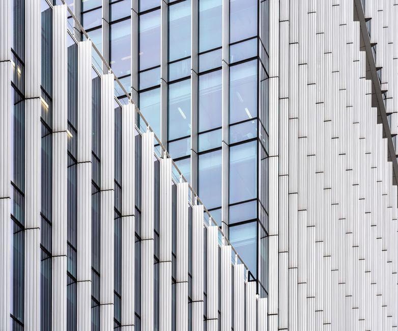 A modern building facade seen at an angle: vertical white metal fins stepping across a blue-tinted glass curtain wall, a diagonal edge cutting the frame