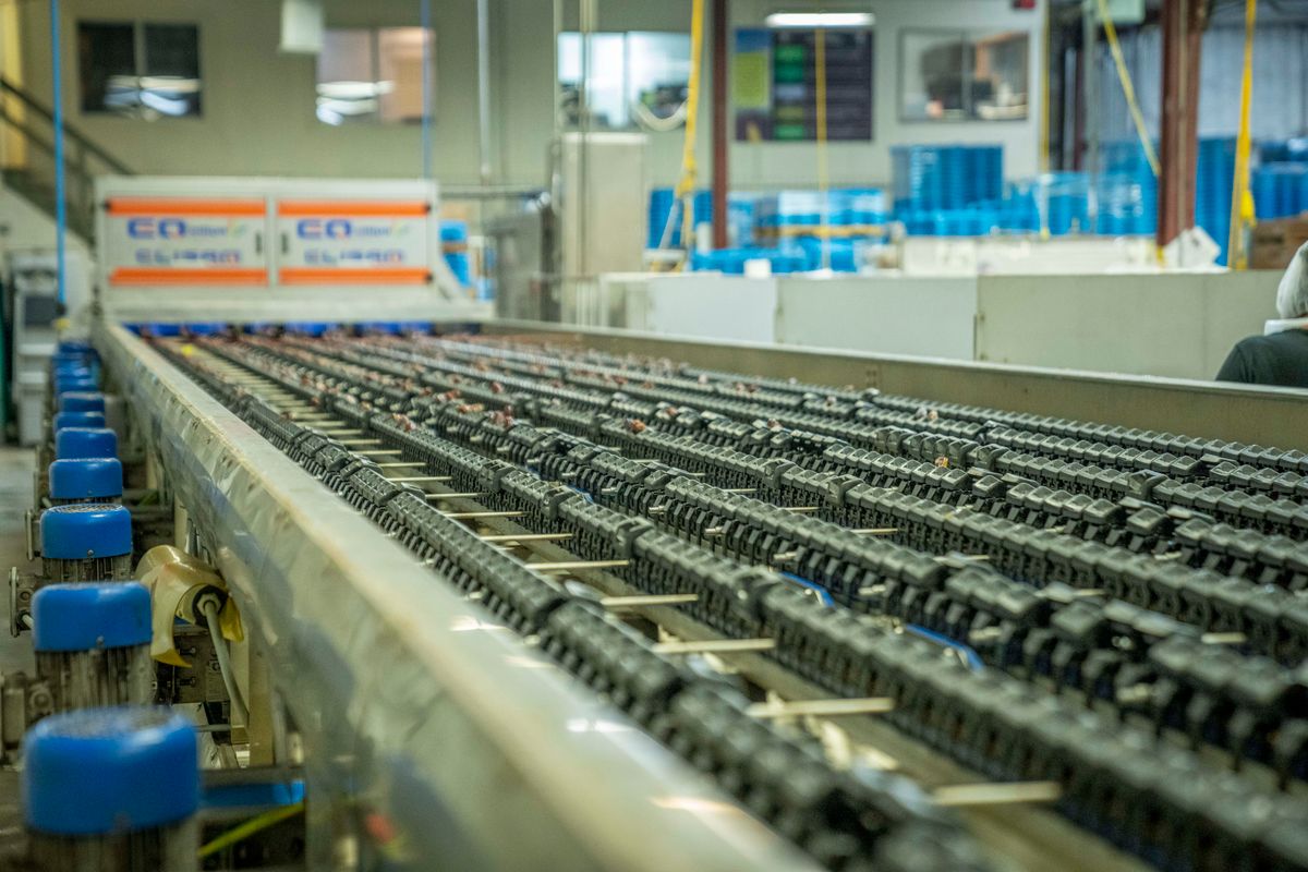 Una línea transportadora industrial que recorre una fábrica, con hileras de piezas metálicas oscuras sobre ella, elementos con tapa azul a la izquierda y maquinaria desenfocada al fondo. Un canal nightly es esto y no una cuenta atrás: produce, y sigue produciendo.