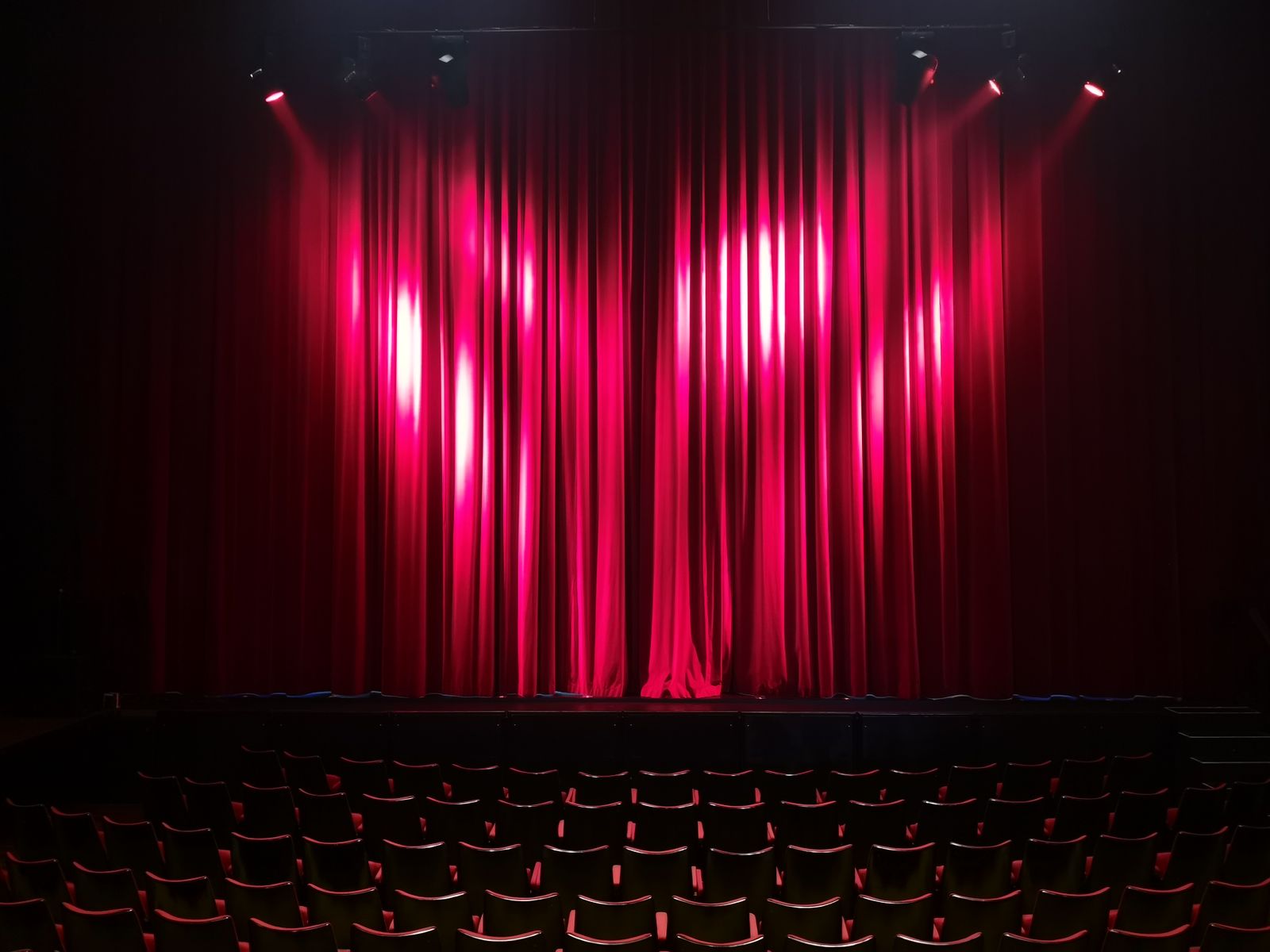 Un théâtre vide avec un rideau rouge éclairé par des projecteurs, des rangées de sièges vides au premier plan